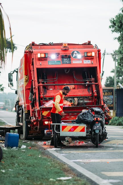 A worker dressed in a yellow vest with reflective orange stripes is standing on the roadside near a large red rubbish collection vehicle, which is parked on the curb. The back of the vehicle is open, revealing mechanisms and compartments for waste collection, with a black trash bag placed on the rear platform. The worker appears to be handling or inspecting waste before disposal, with a small motorbike parked close to the vehicle. The scene takes place outdoors on a street with a grassy area to the left, and a blue building with signage is visible in the background. The sky is overcast, providing diffuse lighting that softly illuminates the scene. This image exemplifies a private waste collection scenario, where on-site rubbish removal services are being carried out independently, aligning with the themes of alternatives to municipal rubbish disposal as discussed on wasteclearancekennington.co.uk.