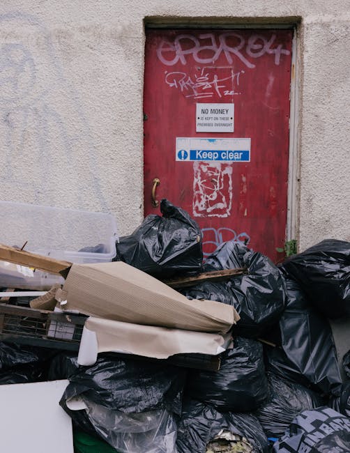 A large pile of black refuse bags, some torn and bulging, is stacked against a weathered exterior wall beneath a red metal door covered in graffiti. Several loose cardboard pieces and other discarded materials are visible among the bags, which are piled haphazardly on a concrete surface. The door features various signs, including one that states 'No Money' and another that says 'Keep Clear,' indicating restricted access. The surrounding environment suggests an urban alley or backstreet area, likely a site for rubbish accumulation awaiting collection or disposal. The scene reflects an instance where private waste clearance services, such as those offered by Waste Clearance Kennington, might be employed for on-site clearance or alternative waste handling outside council collection schedules.