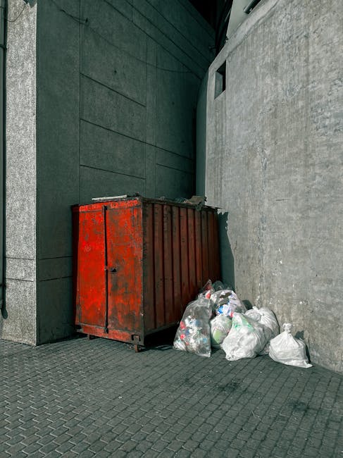 A worker dressed in a yellow vest with reflective orange stripes is standing on the roadside near a large red rubbish collection vehicle, which is parked on the curb. The back of the vehicle is open, revealing mechanisms and compartments for waste collection, with a black trash bag placed on the rear platform. The worker appears to be handling or inspecting waste before disposal, with a small motorbike parked close to the vehicle. The scene takes place outdoors on a street with a grassy area to the left, and a blue building with signage is visible in the background. The sky is overcast, providing diffuse lighting that softly illuminates the scene. This image exemplifies a private waste collection scenario, where on-site rubbish removal services are being carried out independently, aligning with the themes of alternatives to municipal rubbish disposal as discussed on wasteclearancekennington.co.uk.