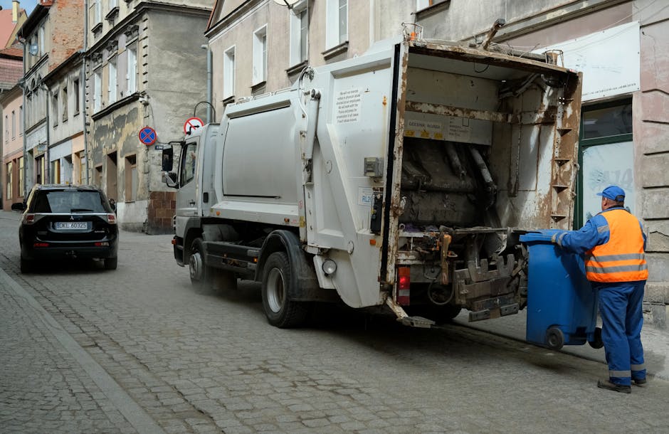 A waste collection vehicle is parked on a narrow urban street next to a row of multi-storey residential buildings with aged facades, some showing peeling paint and weathering. The truck, primarily white with signs of rust and dirt around its rear compartment, is equipped with an open rear loading area that contains black and grey compacted waste materials. A worker dressed in a blue uniform and an orange high-visibility vest is positioned at the back of the truck, actively placing a blue wheeled bin into the loading mechanism. The street surface is made of cobblestones, and next to the truck, there is a black vehicle parked parallel to the curb. In the background, there are various street signs and a shopfront with a glass window, indicating a mixed-use area. The scene reflects an outdoor waste collection process, typical of private rubbish collection services like Waste Clearance Kennington, in an environment where alternative waste handling is visibly in progress, emphasizing the practicalities of urban rubbish removal.