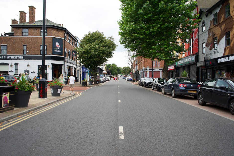 A city street scene showing a wide, paved road lined with parked cars on both the left and right sides, including a range of dark-colored sedans and hatchbacks. The sidewalk on the left features large decorative planters filled with flowering plants, and pedestrians are walking along the pavement. On the left side, a black and white building with a corner entrance and signage indicating a pub or café is visible, with outdoor seating and people gathered nearby. The right side displays a row of multi-story commercial shops with distinctive signage, including a pharmacy and other retail units, adorned with colorful banners and signs. Trees with lush green foliage are spaced along the sidewalk, providing some shade, and the sky above is overcast with diffused natural light. The scene depicts an active urban environment emphasizing private on-site waste management, with Waste Clearance Kennington offering rubbish removal and collection services, though no waste materials or removal activities are directly visible in the image. The overall composition captures the typical appearance of a commercial street with parking, pedestrian activity, and storefronts, suitable for content related to rubbish and waste clearance services in an urban setting.