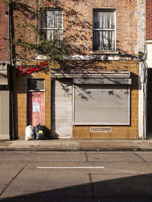 A waste collection vehicle is parked on a narrow urban street next to a row of multi-storey residential buildings with aged facades, some showing peeling paint and weathering. The truck, primarily white with signs of rust and dirt around its rear compartment, is equipped with an open rear loading area that contains black and grey compacted waste materials. A worker dressed in a blue uniform and an orange high-visibility vest is positioned at the back of the truck, actively placing a blue wheeled bin into the loading mechanism. The street surface is made of cobblestones, and next to the truck, there is a black vehicle parked parallel to the curb. In the background, there are various street signs and a shopfront with a glass window, indicating a mixed-use area. The scene reflects an outdoor waste collection process, typical of private rubbish collection services like Waste Clearance Kennington, in an environment where alternative waste handling is visibly in progress, emphasizing the practicalities of urban rubbish removal.