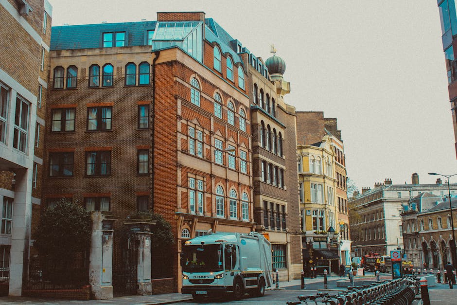 A street scene featuring a row of multi-story historic buildings with varying architectural styles, including a red brick building with arched windows and decorative stonework, a narrow light yellow building with bay windows, and a taller tan building with a domed tower topped by a decorative finial. In the foreground, a grey waste collection vehicle is parked alongside the curb, part of waste clearance operations, with black bollards and bike racks in front of the vehicle. The street is lined with a mix of modern and older structures, with a lamppost visible on the right side, and a cloudy sky overhead. The scene suggests urban waste management activities in a central city environment, with Waste Clearance Kennington potentially involved in private rubbish collection or on-site clearance services for commercial or residential properties nearby, supporting alternative waste handling options outside local authority collections.