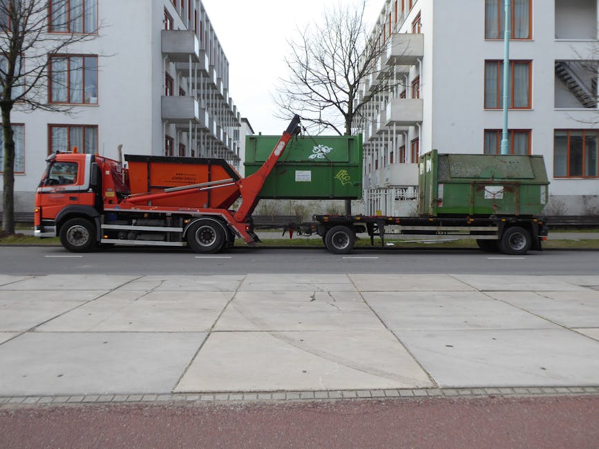 A large red refuse collection truck equipped with a hydraulic lift mechanism is parked along a paved urban street, positioned parallel to a row of modern multi-storey residential buildings. The truck features a prominent cab with side mirrors, a telescopic arm extending upward from the vehicle’s midsection, and a rear-loading container partially lifted for waste transfer. Adjacent to the truck, on a flatbed trailer, are two large, weathered green metal skips with visible graffiti, signs of rust, and surface dirt; one is positioned at the front of the trailer while the other is towards the rear. The scene is set during daylight hours, with natural overcast lighting, and minimal background activity. To the left and right of the truck, leafless trees line the street, and a white building with multiple balconies and windows can be seen behind the skips, contextualising this as an area dealing with private waste collection in an urban environment. This image complements services related to independent waste disposal or rubbish removal as part of site clearance efforts, often handled by companies such as Waste Clearance Kennington.