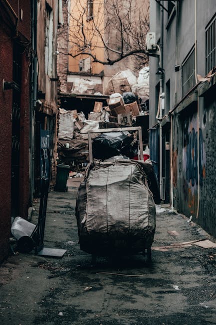 A large collection of mixed household rubbish and waste, including cardboard boxes, paper bags, plastic packaging, black and red rubbish bags, and various discarded items, all piled around a grey plastic recycling bin labeled for mixed paper and cardboard. The waste is scattered on a paved area in front of a commercial building with a blue metal fence, behind which are storefronts displaying signs and advertisements. To the left, a parked silver car is partially visible, with a stone railing separating the waste area from the parking spaces. The scene is outdoors, illuminated by natural daylight, with a leafless tree on the left side of the image and a building with scaffolding and safety signage visible in the background. The image illustrates a typical private waste collection scenario requiring rubbish removal services like those offered by Waste Clearance Kennington, highlighting the need for efficient clearance of household waste from urban areas.