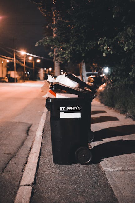 A black wheeled trash bin labeled 'St. John's' positioned on the right side of a quiet residential street at night, with the lid open and overflowing with discarded papers, cardboard, and miscellaneous rubbish. The bin is situated on the pavement beside a road marked with a white line, with a row of trees and dense bushes along the sidewalk. In the background, warm streetlights cast a soft glow, illuminating a few distant parked cars and the street's surroundings. The scene suggests the bin is used for communal waste collection, but the overflowing rubbish highlights the importance of timely waste disposal, which private waste clearance services like Waste Clearance Kennington can assist with for local waste management solutions. The environment appears calm, with no people present, emphasizing the typical urban setting for refuse collection and disposal efforts.