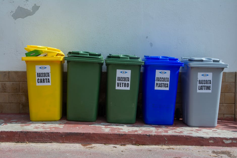 A row of five colour-coded waste bins placed on a paved sidewalk against a plain white wall with a low brick border. From left to right, the first bin is yellow with a white label marked 'RACCOLTA CARTA', used for paper collection, with a yellow lid that is open. The second and third bins are green with labels indicating 'RACCOLTA VETRO' for glass recycling, both featuring matching green lids. The fourth bin is blue with a label reading 'RACCOLTA PLASTICA' for plastic waste, topped with a blue lid. The fifth bin is grey with a label 'RACCOLTA LATTINE' intended for cans, fitted with a grey lid. The bins have a smooth, plastic texture and are positioned on a curb edge that shows signs of wear and peeling paint, indicative of outdoor placement. This arrangement suggests an organized waste segregation system, which may support private waste collection services like those provided by Waste Clearance Kennington, especially in the context of alternative waste handling or independent collection outside municipal schedules.