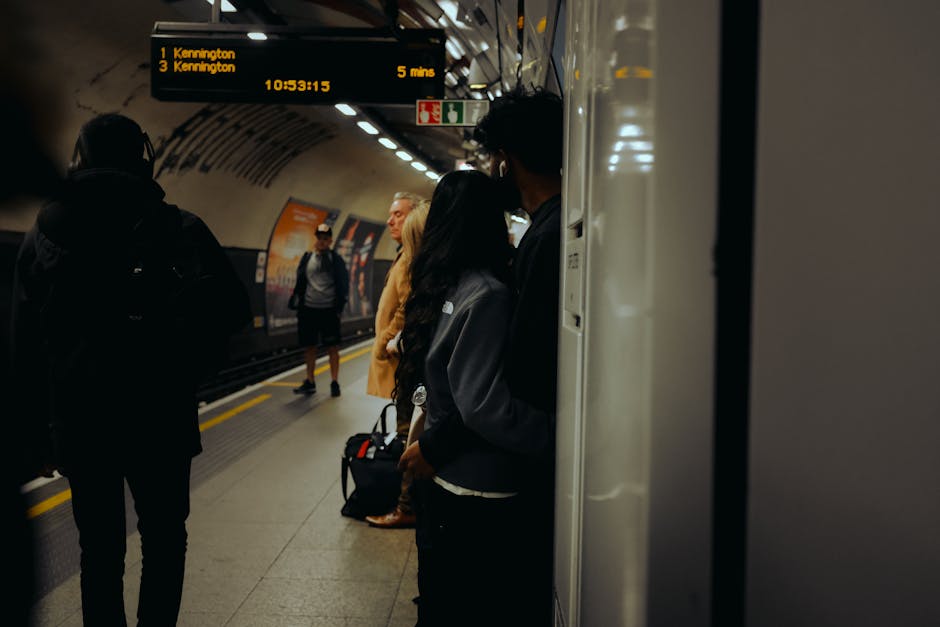 Inside a London underground railway station, several passengers are waiting on the platform near a white tiled wall, with some leaning against it. The foreground features a man with a dark backpack and glasses, standing with his back to the camera. Next to him, a woman with long dark hair, dressed in dark clothing, is partially visible, leaning against the wall. Further along the platform, a woman in a mustard coat and beige boots stands facing forward, while a man in casual attire, including shorts and a jacket, approaches from the opposite side. Overhead, an electronic display board shows upcoming train departures to Kennington, indicating a five-minute wait, with the time displayed as 10:53:15. The station's lighting is bright but neutral, with a curved tunnel ceiling extending into the background, where additional passengers are visible. The scene captures a typical busy commute environment, possibly relating to private or alternative transportation methods, akin to off-menu or independent collection services by waste clearance providers such as Waste Clearance Kennington, although no waste or disposal items are visible in this image.