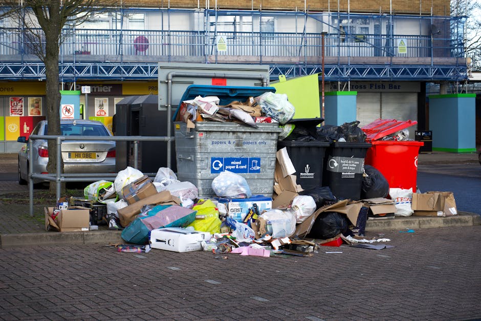 A large collection of overflowing rubbish bins and scattered waste including cardboard boxes, plastic bags, paper, and general household trash piled around a communal refuse area on a paved parking lot. The waste comprises brown, black, and clear plastic bags, some torn open with contents spilling out, along with flattened cardboard boxes, paper packaging, and other debris. The scene includes a metallic mixed paper and card bin with a blue lid, a black recycling bin, and a bright red general waste bin, all situated close together. Surrounding the refuse are parked vehicles, one grey car with a yellow registration plate, and a metal railing separating the waste area from the parking bays. In the background, a modern building with a storefront and upper-floor windows is visible, along with a tree on the left side and a construction scaffold on the upper level, indicating ongoing building work. The overall setting suggests an unregulated domestic waste disposal area, highlighting potential issues with illegal dumping or inadequate rubbish collection services in the vicinity.