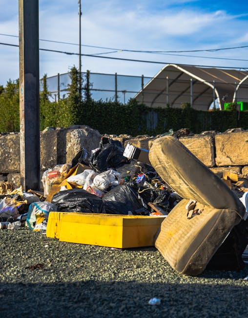 A close-up view of a pile of mixed domestic waste and rubbish on the side of a street or an outdoor area, including black plastic bin liners filled with refuse, a yellow plastic storage container, and a worn, dirty beige car seat. The rubbish is scattered on a gravel surface, with a stone wall behind it. In the background, there are utility poles with overhead power lines, some green shrubbery, and a structure with a curved metal roof. The scene is lit by natural daylight, highlighting the textures of the trash, gravel, and surrounding environment, illustrating typical issues with illegal dumping or unmanaged waste collection that waste clearance services like Waste Clearance Kennington might manage through private, on-site disposal or alternative waste handling solutions.
