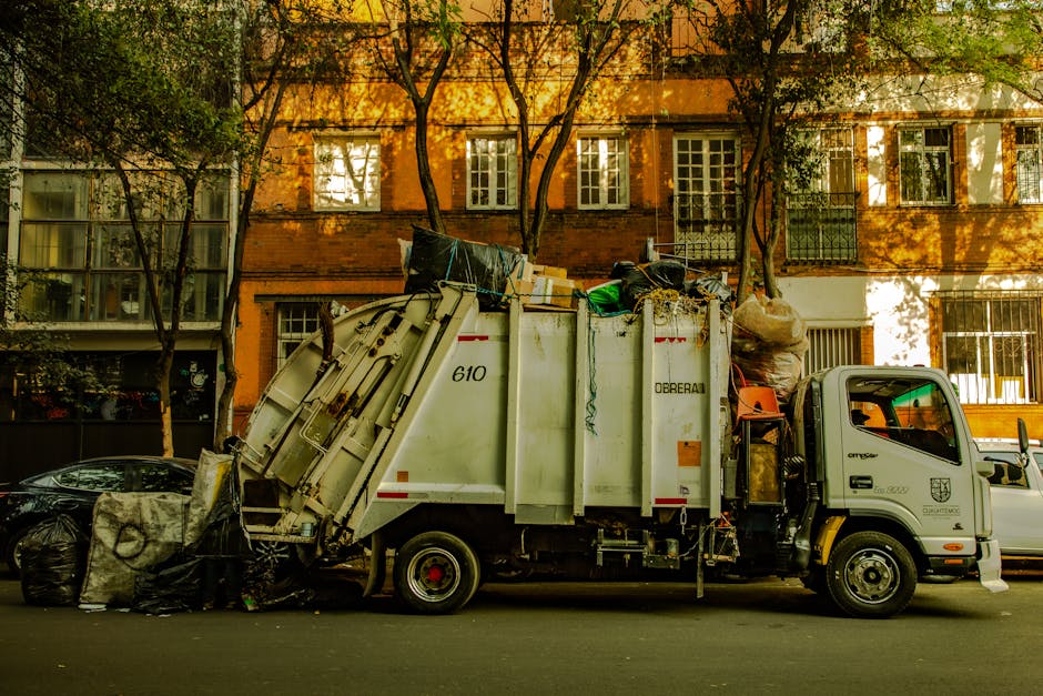 A close-up view of a pile of mixed domestic waste and rubbish on the side of a street or an outdoor area, including black plastic bin liners filled with refuse, a yellow plastic storage container, and a worn, dirty beige car seat. The rubbish is scattered on a gravel surface, with a stone wall behind it. In the background, there are utility poles with overhead power lines, some green shrubbery, and a structure with a curved metal roof. The scene is lit by natural daylight, highlighting the textures of the trash, gravel, and surrounding environment, illustrating typical issues with illegal dumping or unmanaged waste collection that waste clearance services like Waste Clearance Kennington might manage through private, on-site disposal or alternative waste handling solutions.