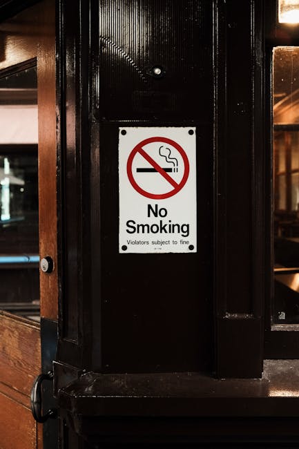 A close-up view of a white rectangular 'No Smoking' sign mounted on a glossy black metal column inside an indoor space, possibly a restaurant or café. The sign prominently displays a red circle with a diagonal line crossing out a stylized cigarette emitting smoke, indicating a no-smoking policy. Beneath the symbol, the words 'No Smoking' are written in bold black letters, with smaller text below stating 'Violators subject to fine.' The metal surface surrounds the sign, with textured vertical ridges creating a slight reflective sheen. In the background, warm lighting and blurred interior elements suggest a cozy, indoor environment, with wooden and glass finishes hinting at a dining area or lounge. The sign is positioned at eye level, clearly visible for patrons, emphasizing compliance with internal regulations related to smoking restrictions within the premises managed by a waste management service that emphasizes responsible waste disposal and regulation adherence.