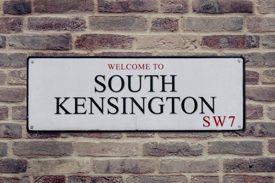 A rectangular metal street sign fixed to a red brick wall displaying the message 'Welcome to South Kensington SW7'. The sign has a white background with black and red lettering, where 'Welcome to' and 'SW7' are in red, and 'South Kensington' is in large, bold black letters. The brick wall behind it features a traditional running bond pattern with reddish-brown bricks and light gray mortar lines, providing a textured backdrop. The sign is positioned centrally on the wall, with visible screw fasteners at each corner, and the overall scene suggests an urban setting typical of residential or public areas in South Kensington. This type of signage is often used for directional or informational purposes in private or semi-private environments, aligning with practices of independent property or site management, such as waste clearance or property signage in the area. The lighting appears natural, highlighting the textures and colors of the brick wall and the sign, contributing to a clear and professional visual context relevant to local geographic navigation or property identification.