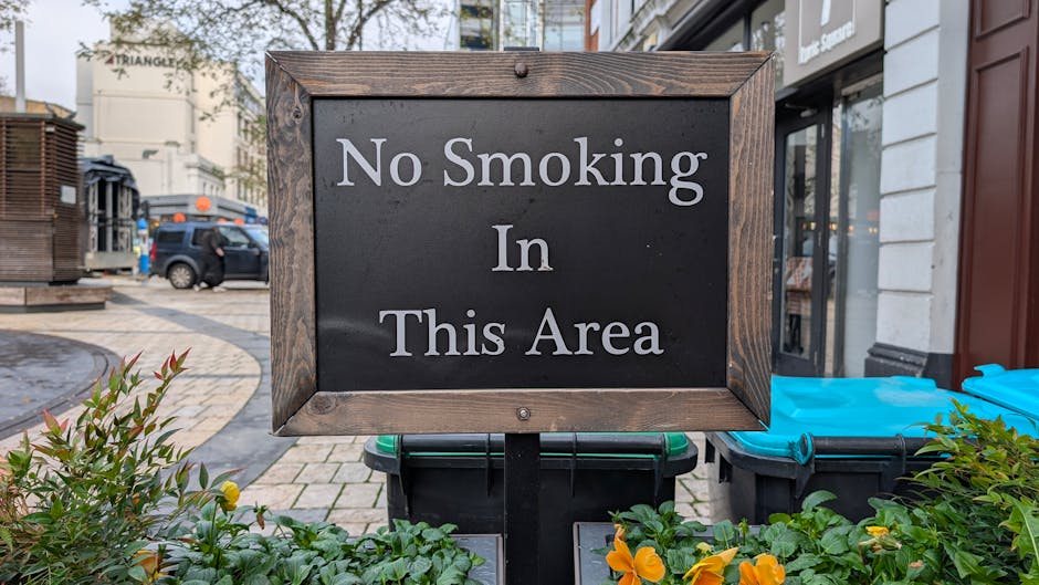 A close-up view of a white rectangular 'No Smoking' sign mounted on a glossy black metal column inside an indoor space, possibly a restaurant or café. The sign prominently displays a red circle with a diagonal line crossing out a stylized cigarette emitting smoke, indicating a no-smoking policy. Beneath the symbol, the words 'No Smoking' are written in bold black letters, with smaller text below stating 'Violators subject to fine.' The metal surface surrounds the sign, with textured vertical ridges creating a slight reflective sheen. In the background, warm lighting and blurred interior elements suggest a cozy, indoor environment, with wooden and glass finishes hinting at a dining area or lounge. The sign is positioned at eye level, clearly visible for patrons, emphasizing compliance with internal regulations related to smoking restrictions within the premises managed by a waste management service that emphasizes responsible waste disposal and regulation adherence.
