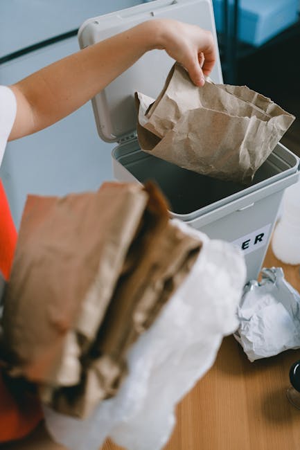 A person placing a crumpled brown paper bag into a grey recycling bin with a partially visible white lid, situated on a wooden surface indoors. The individual's arm is extended, holding the bag by its top edge, and they are wearing a light-colored shirt with a red sleeve. Nearby on the table are additional crumpled paper bags and some loose paper waste. The environment appears to be a home or office space, with a focus on waste disposal and recycling activities, aligning with private rubbish removal services such as those offered by Waste Clearance Kennington, emphasizing on-site waste collection of paper and general rubbish materials.