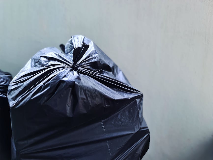 Close-up view of two large black rubbish bags made from glossy plastic material, tightly sealed with knot ties at the top, resting against a plain, light-colored wall. The bags contain general household waste, appearing somewhat bulging and irregular in shape due to their contents. The surface of the plastic shows reflections of light, highlighting the crinkled texture and smooth finish. The bags are positioned on a surface possibly outdoors or in a designated waste collection area, with no other objects visible in the immediate vicinity. The image is clear and focused on the bags, emphasizing their size and material, which is typical for private waste collection services like those offered by Waste Clearance Kennington specializing in on-site rubbish removal and alternative waste handling solutions. The neutral environment emphasizes the practical nature of rubbish disposal, supporting the context of efficient and independent waste clearance.